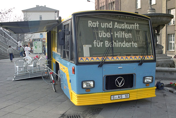 Ein großer, blau-gelber Reisebus steht auf der Straße. Er ist zu einem Infostand umfunktioniert und hat beide Seitenteile ausgeklappt. Auf der Windschutzscheibe steht Rat und Auskunft über Hilfen für Behinderte.